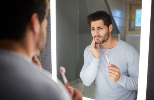 Wincing man in bathroom holding bloody toothbrush in one hand and touching his face with the other