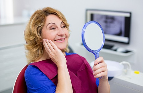 Older female patient checking smile in mirror after receiving implant dentures