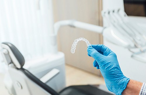 Gloved hand holding Invisalign clear aligner in front of dental chair